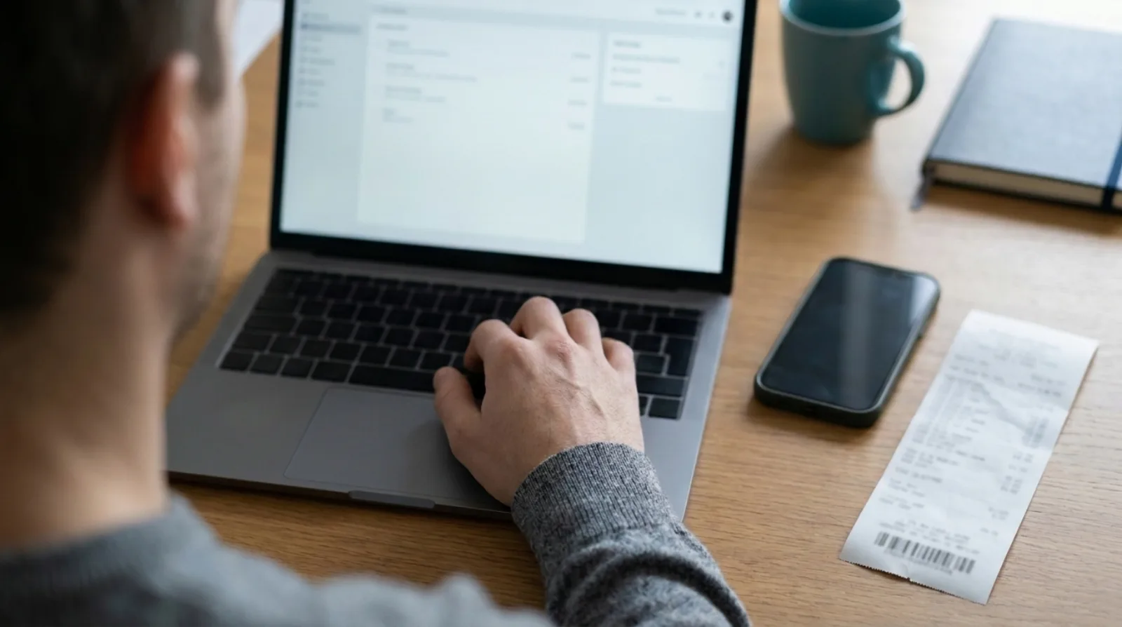 A person seen from behind typing on a laptop showing a blurred spreadsheet, with a dark phone and a long narrow till receipt lying on the wooden desk beside it.